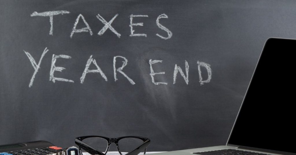 Taxes Year End Written on A Chalkboard With A Laptop, glasses, watch, and calculator on a desk. | End of Year Accounting